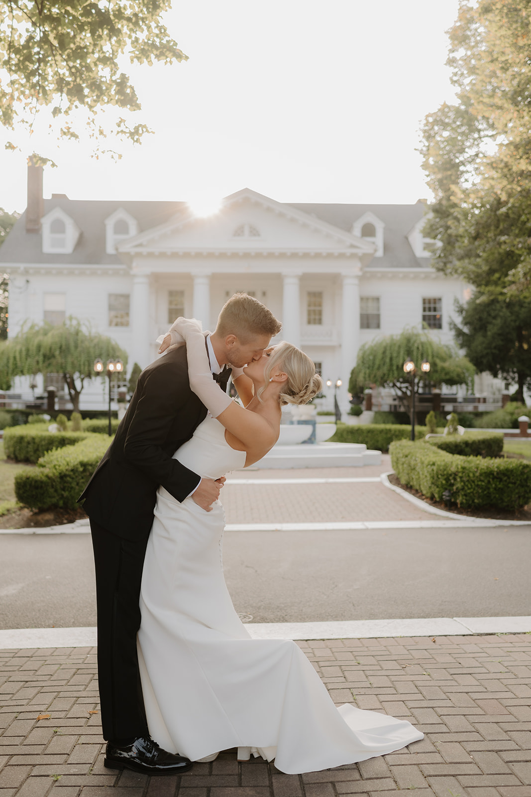 Groom dips and kisses the bride on a brick path, with the historic white mansion shining behind them.